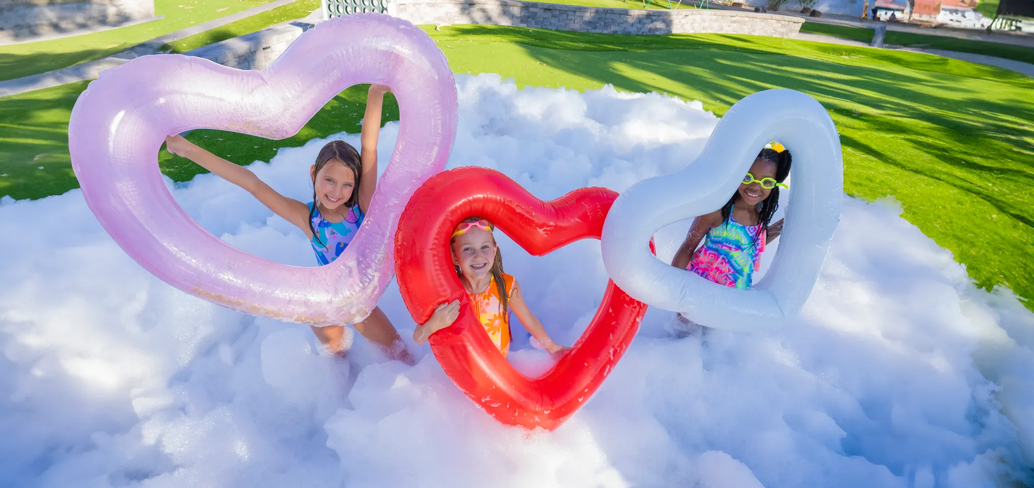 Three girls holding colorful heart-shaped inflatables while standing in foam at a Beverly foam party