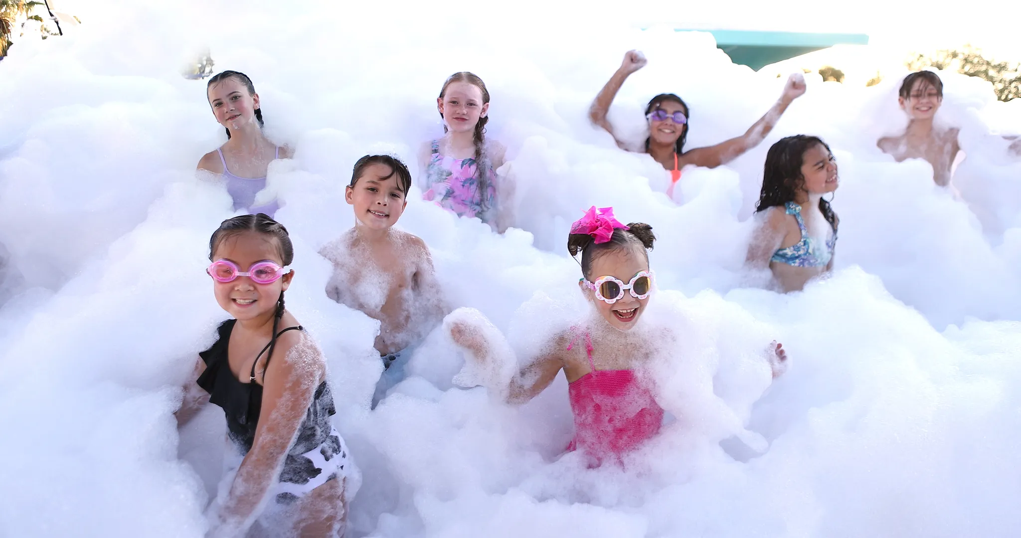 Group of eight kids smiling and cheering while chest-deep in foam at a foam party