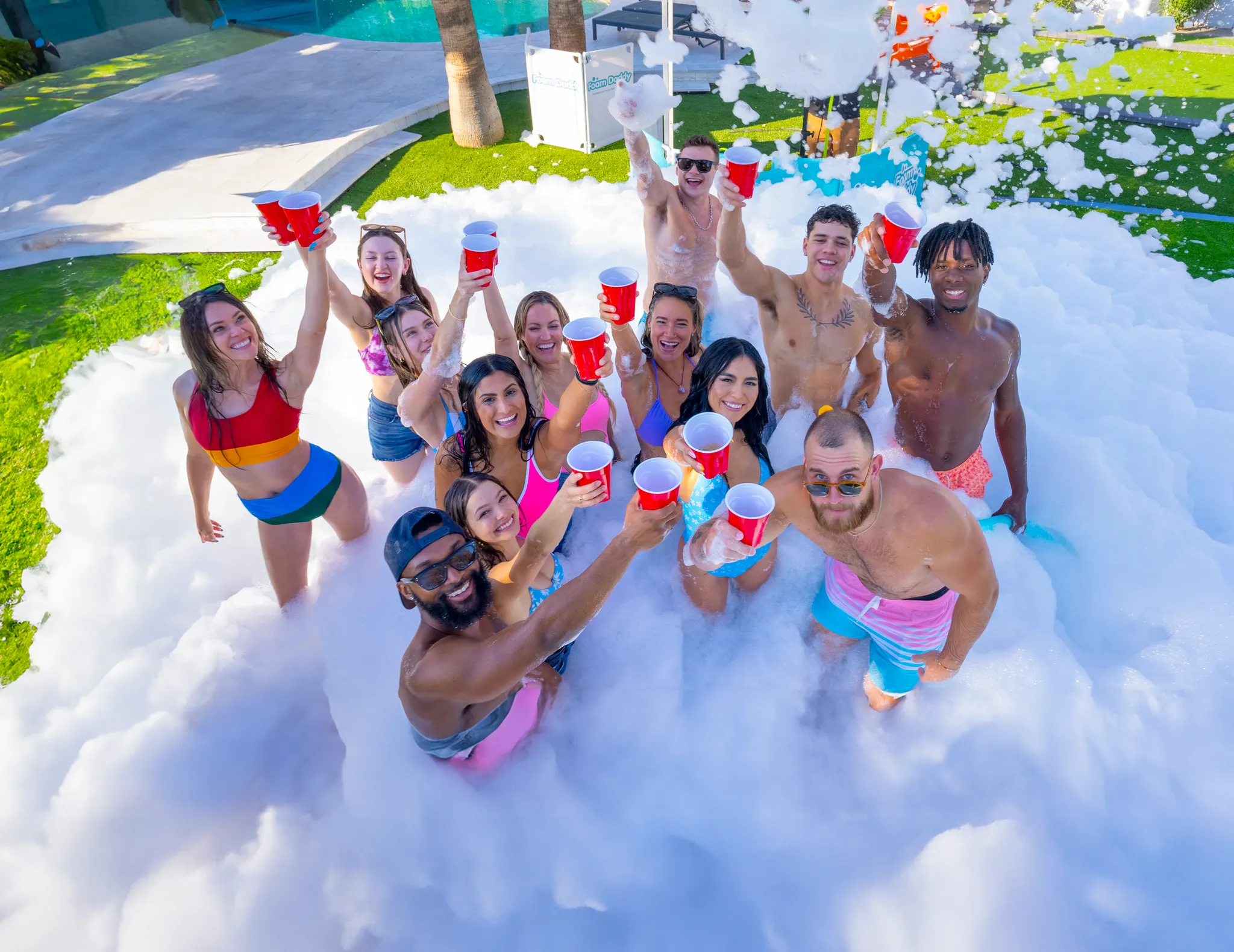 Group of young adults raising red cups and cheering while standing in foam at an outdoor community foam party