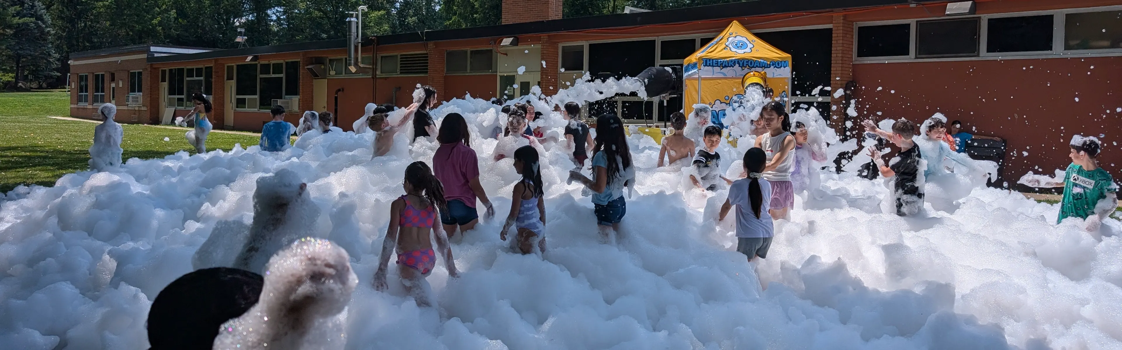 A large group of kids playing in a wide field of foam beside the Party Foam tent at a sunny school event