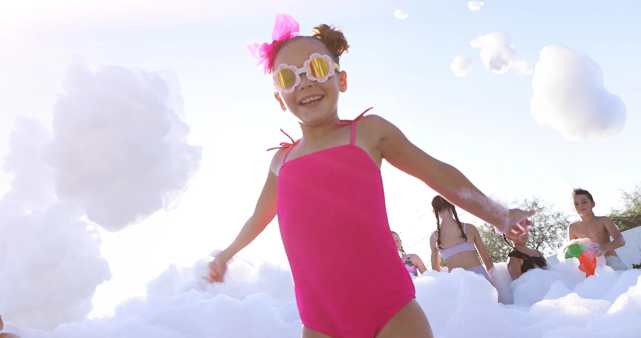 Girl in a bright pink swimsuit and flower sunglasses smiling while playing in foam at a birthday foam party