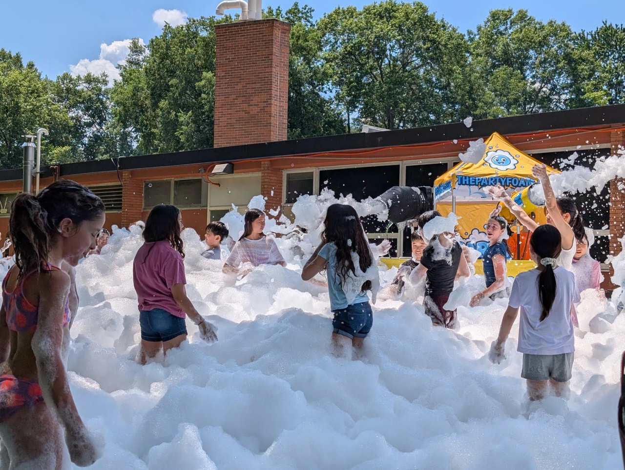 Kids playing across a field of deep foam beside the Party Foam cannon and branded tent at a New Jersey school event